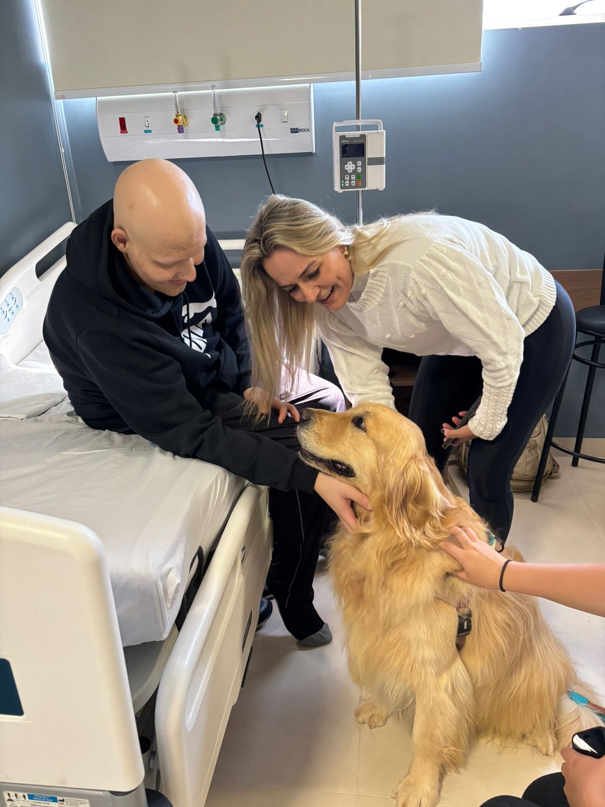 A bald man sits on a hospital bed, smiling at a golden retriever being petted by a woman. Another person is nearby, interacting with the dog.