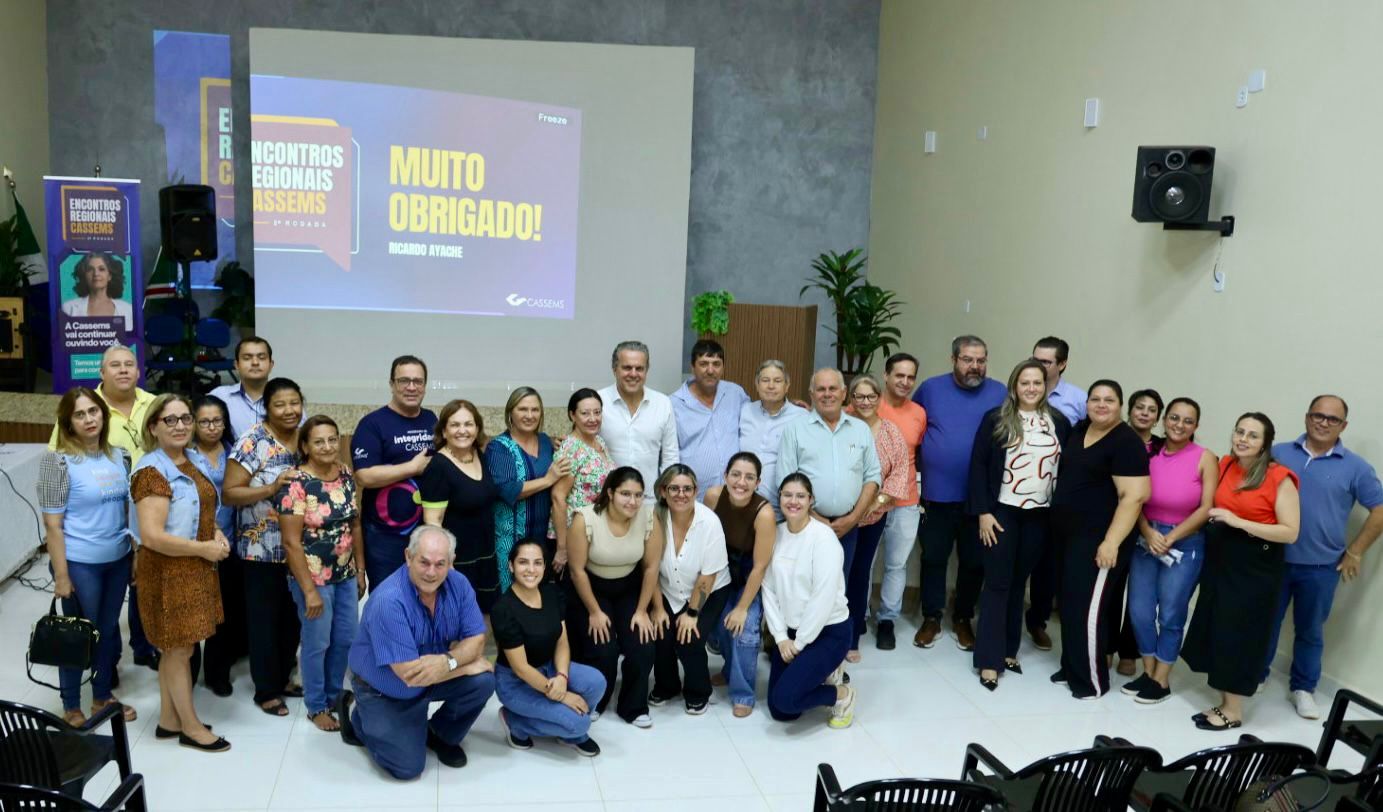 A group of people poses together in a conference room, with a presentation screen in the background displaying "MUITO OBRIGADO!"