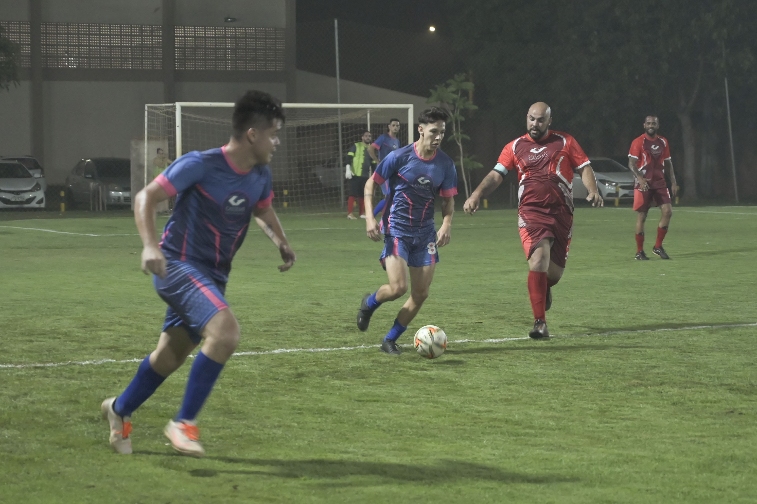 Players in colorful jerseys are engaged in a soccer match on a well-maintained grass field at night.