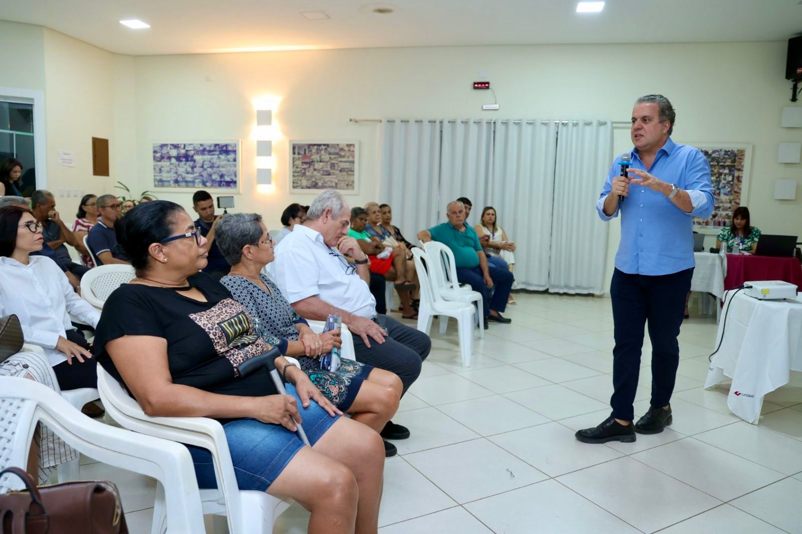 A speaker addresses an audience seated in a well-lit room, with attendees engaged and listening attentively.