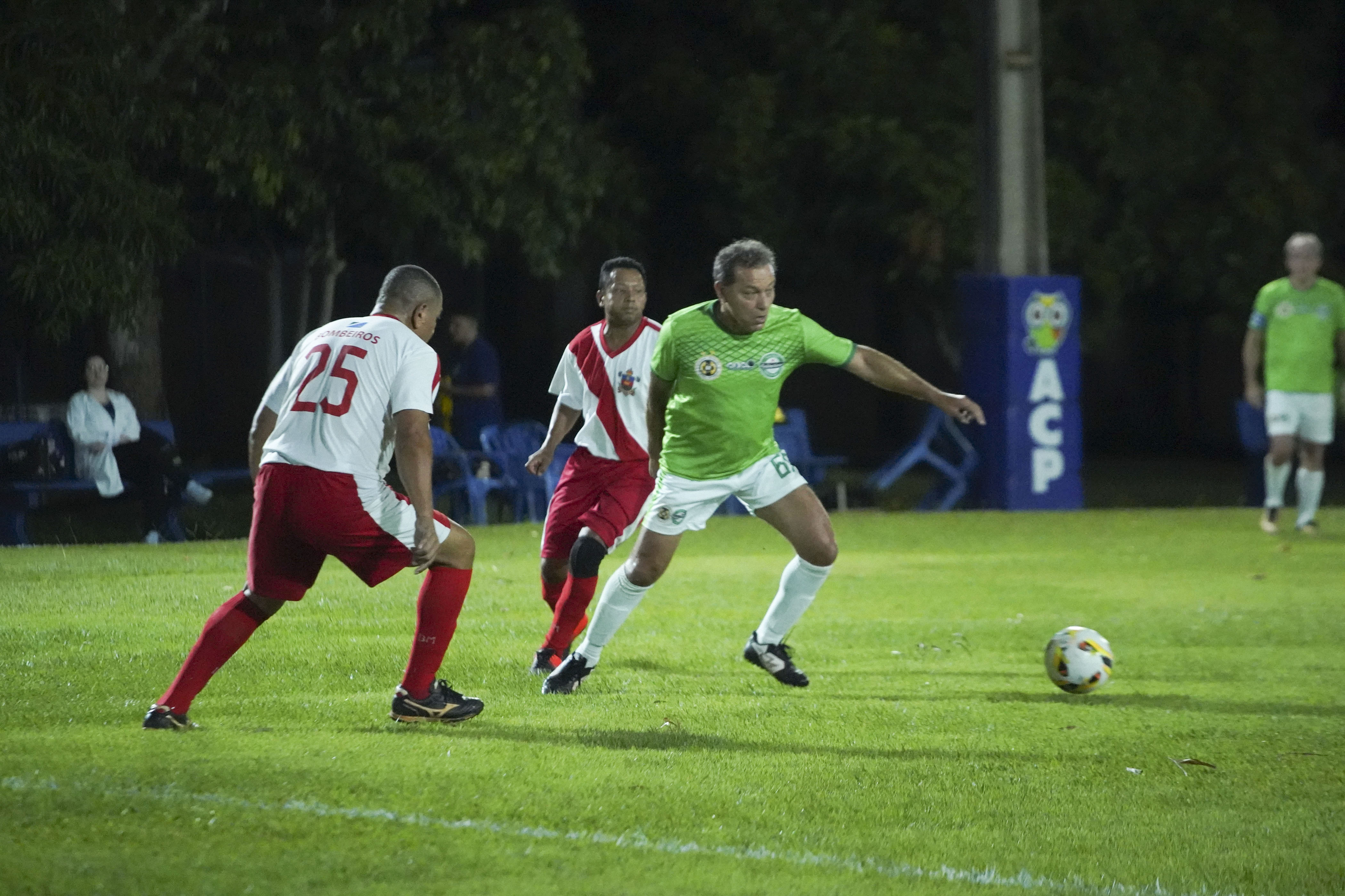 A soccer match in progress at night, featuring players in green and red uniforms competing for the ball on a grassy field.