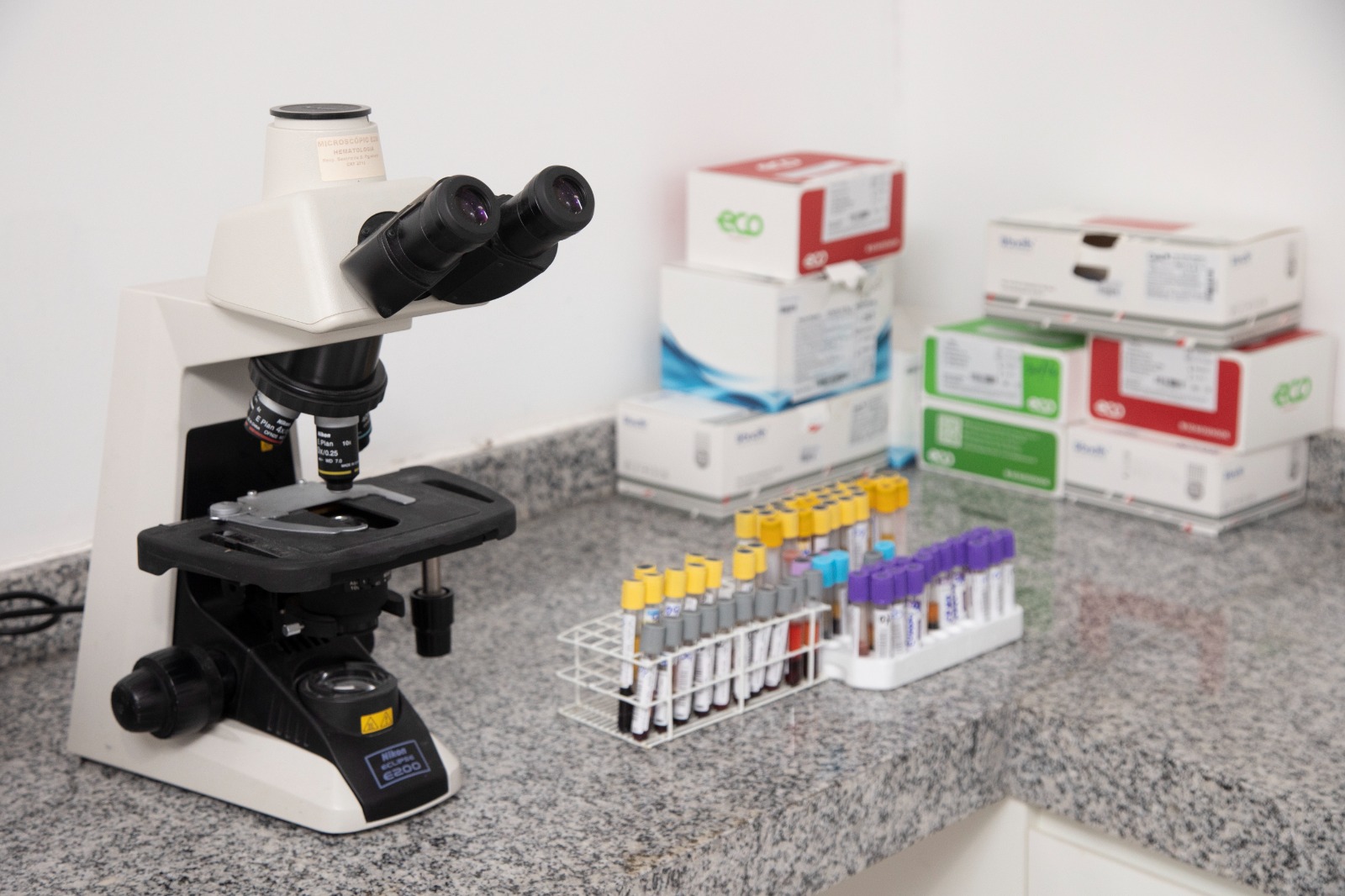 A laboratory setup featuring a microscope and a rack of test tubes, with various boxes in the background.