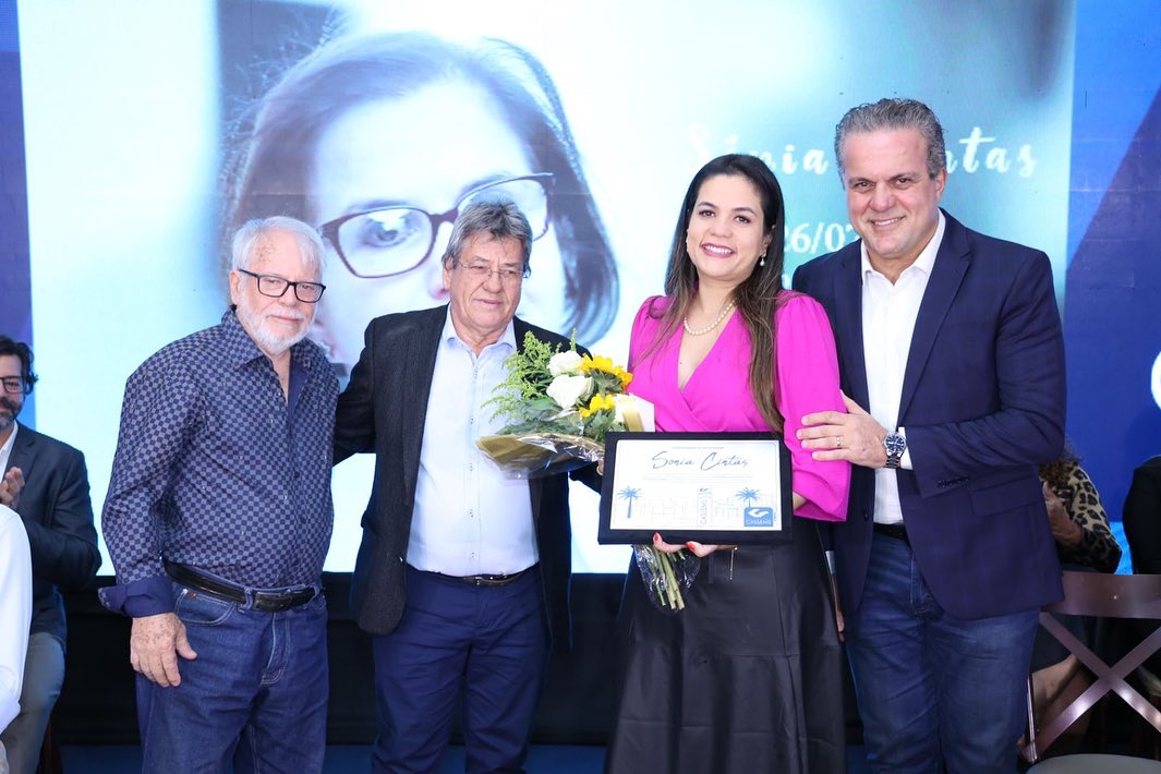 Four people posing, one woman holding a certificate and flowers, with a large portrait in the background.