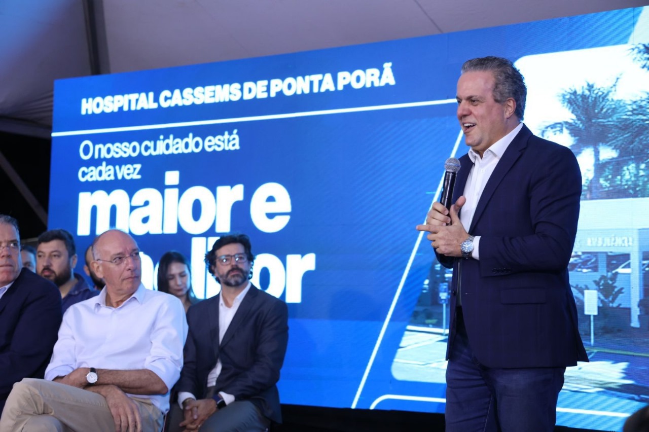 A man in a suit speaks into a microphone on stage, with several seated individuals behind him. A large screen displays text about Hospital Cassems de Ponta Porã.